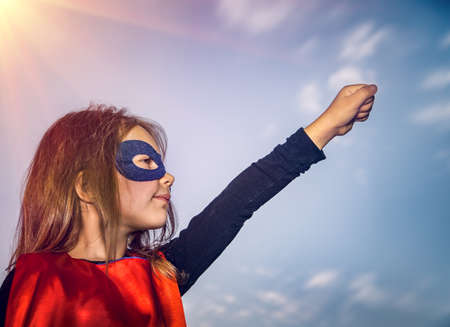 Profile Photo of a Nice Little Girl Dressed as Supergirl with Raised up Hand Outdoors. Happy Joy Time. Conceptual Photo of Feminisms and Women Rights.の写真素材