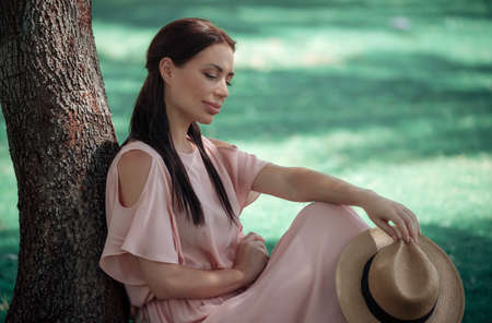 Beautiful Young Female Spending Leisure Time in the Park. Resting near the Tree with Strow Sun Hat in Hand. Happy Life.の写真素材