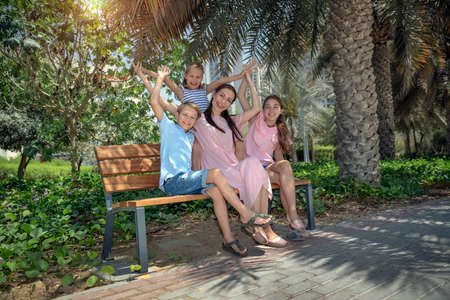 Happy Mother and Her Three Precious Kids Sitting on the Bench in the Park. Big Family Having a Good Time in City Park on Summer Day. Love Concept.の写真素材