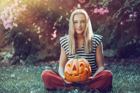 Pretty Woman Sitting in Backyard with Big Carved Pumpkin Head. Traditional Halloween Decoration. Autumn Holiday.の写真素材