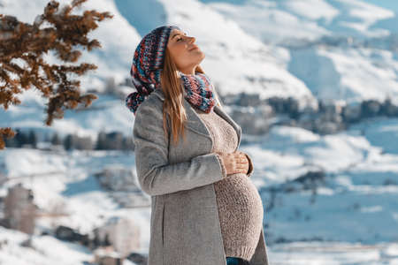 Nice Pregnant Woman With Closed Eyes of Pleasure Enjoying Bright Sun Light in Snowy Mountains. Spending Pregnancy Time in Winter Mountainous Resort.の写真素材