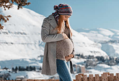 Nice Pregnant Woman with Love Looking at her Tummy. Enjoying Bright Sun Light in Snowy Mountains. Spending Pregnancy Time in Winter Mountainous Resortの写真素材