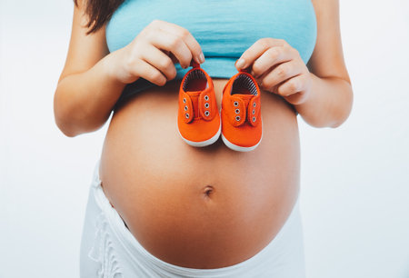 Closeup Photo of a Tummy of Pregnant Woman with Tiny Red Baby Boots Isolated on White Background. New Life Concept.の写真素材