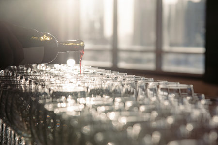 a waiter pouring red wine from bottle to wineglass that set up for dinner reception party in evening time. photo has some flare effect from back light of sun outside of windows behind.の写真素材
