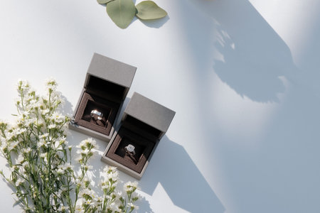 The wedding rings are in boxes on a corner of a white table, decorated with dried flowers, basking in the morning sunlight and shadow in a blank space.