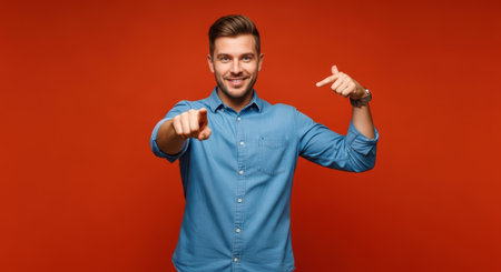 Confident young caucasian male pointing and smiling in blue shirt against orange backgroundの素材
