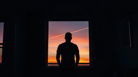 Male silhouette against vibrant sunset viewed through window.の素材