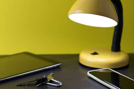 nightstand detail concept with tablet, smart phone, keys and a lighted lampの写真素材
