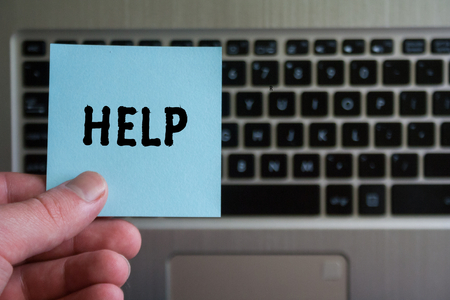 word HELP on sticky note hold in hand on laptop keyboard background.