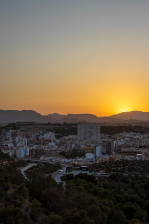 Aerial view of Alicante city at sunset showcasing the skyline against a vibrant orange and blue sky highlighting buildings and lush greenery in the foreground.の写真素材