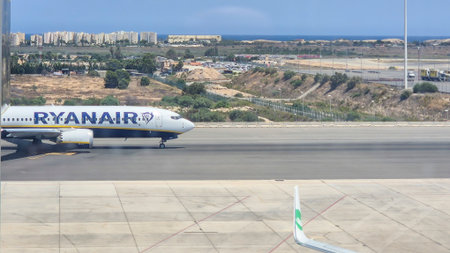 Alicante Spain - July 17 2025: A Ryanair airplane taxiing at the Alicante Airport showing the runway and distant coastline with modern buildings visible.のeditorial素材