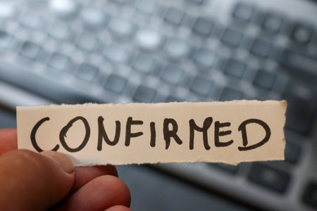 A hand holds a piece of paper with the word CONFIRMED written on it set against a blurred background...
