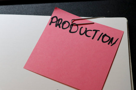 A pink sticky note labeled PRODUCTION is attached to a blank paper using a paperclip symbolizing organization and planning in a workspace setting.の写真素材