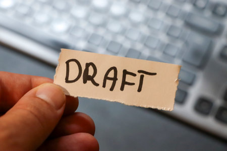 A close-up of a hand holding a torn paper label marked DRAFT against a blurred keyboard background. This image captures the essence of document editing and workflow processes.の写真素材