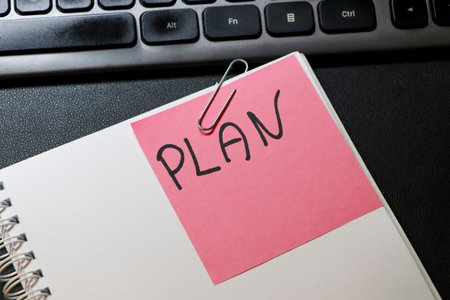 Close-up of a notepad with a pink sticky note labeled PLAN secured by a paperclip next to a computer keyboard symbolizing organization and productivity in an office setting.の写真素材