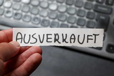A hand holding a sign labeled AUSVERKAUFT meaning sold out in German positioned in front of a blurred computer keyboard. This image captures a moment of unavailability or disappointment.の写真素材