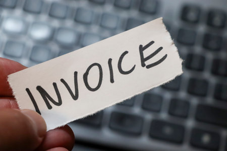A hand holding a torn piece of paper with the word INVOICE written on it set against a blurred computer keyboard background symbolizing financial transactions and business administration.の写真素材