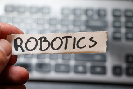 Handwritten word ROBOTICS on a paper strip held above a blurred computer keyboard emphasizing the...