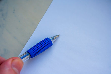 A hand holding a blue pen above a blank sheet of paper ready to write. The clean white background enhances the focus on the pen, ideal for concepts like creativity and planning.の写真素材
