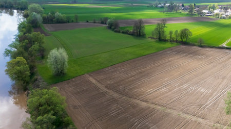 Aerial view of lush green fields alongside a calm river showcasing cultivated lands and trees. The landscape represents agriculture and rural tranquility in springtime.の写真素材