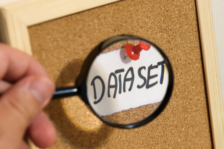 A hand holding a magnifying glass focuses on the word DATASET on a cork board emphasizing research and analysis in data collection and management.の写真素材