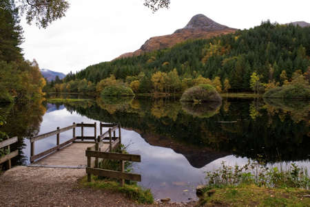 A horizontal image of  a mountain in Glencoe Scotland called the Pap reflecting in the Loch with the surrounding forest and a boat jetty in the foregroundの写真素材