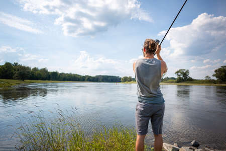 Young fisher on a river bank casting a fishing rod