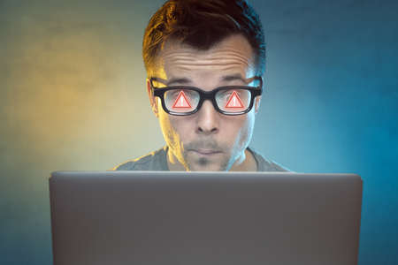 Man in front of a computer with an alert sign reflecting in his glassesの写真素材