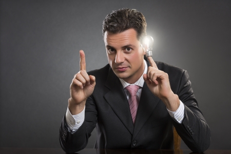 Positive smart consultant pointing up his index finger while holding a bright light bulb at is temple over dark grey background. Smart business strategy concept.の写真素材