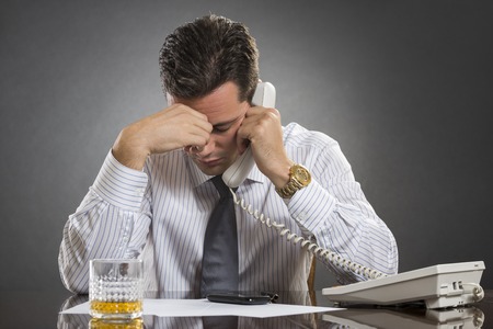 Stressed businessman in white shirt having a bad headache while talking on the telephone against grey , Glass of alcohol on desk.の写真素材