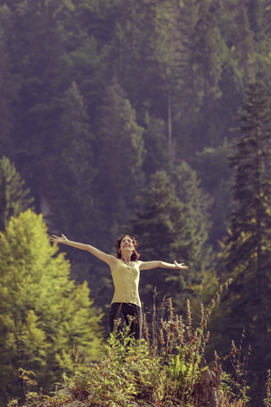 Young delighted woman with arms wide opened, looking up in a gesture of rejoice, freedom and triumph, surrounded by mountain forests.の写真素材