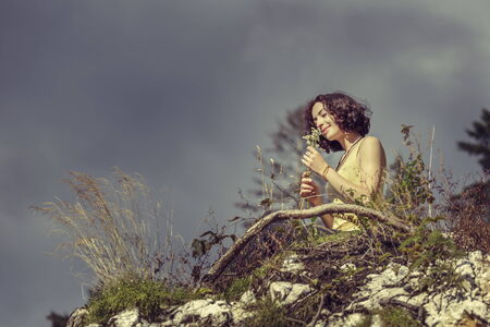 Beautiful happy redhead woman smelling a bouquet of wild mountain flowers while relaxing on top of a rock.の写真素材