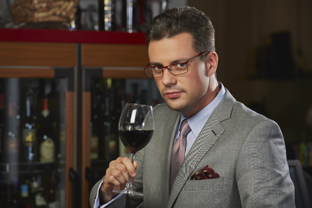 Portrait of sophisticated confident wealthy gentleman in stylish suit toasting with glass of wine against blurred restaurant background.の写真素材