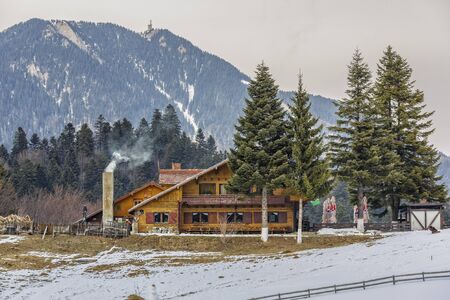 PREDEAL, ROMANIA - FEBRUARY 19, 2015: Rustic wooden Poiana Secuilor chalet with smoking chimney in an overcast winter morning at 1070m altitude. Postavaru mountain (1799 m altitude) in the background.のeditorial素材