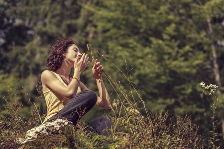 Beautiful peaceful redhead woman gently touching and smelling wild mountain plants while resting on top of a rock.の写真素材