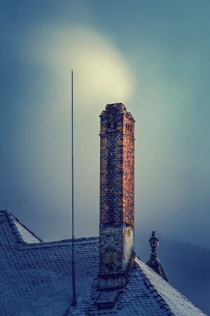 Winter scene with a smoky chimney on a snow covered roof at a gloomy foggy twilight.の写真素材