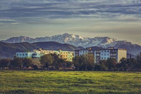 Spring landscape with snowy Bucegi mountains, old block of flats and blooming dandelion fields in the outskirts of Brasov city, Romania.の写真素材