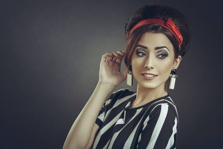 Beautiful smiling woman with hand to ear, wearing striped apparel, red headband and retro updo hairstyle, eavesdrops on fashion rumors, news or trends gossips, over dark background with copy space.の写真素材