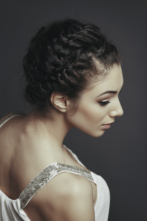 Side face portrait of beautiful female fashion model posing with braided hairdo, wearing white low cut blouse, looking down over grey background.の写真素材