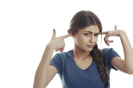 Annoyed pretty young lady with hair braid covers her ears with her fingers when hearing something unpleasant. Pretty woman with disgust grimace, sticks her fingers in her ears refusing to listen.の写真素材