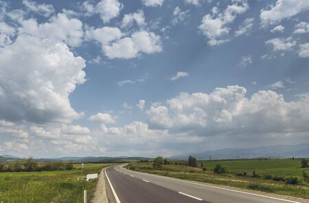 Sunny summer landscape with fluffy clouds in the blue sky and road crossing vast green plains.