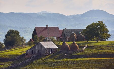 Springtime rural landscape. Traditional Transylvanian house with rustic wooden barn, cowshed, hayricks and green pasture in Brasov county, Transylvania, Romania.の写真素材