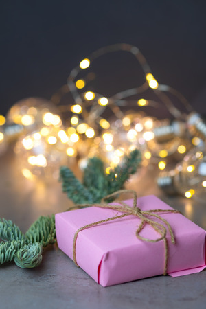 A Christmas present in pink color tied with twine with a fir branch on the background of beautifully blurred garlands. Place for text.の写真素材