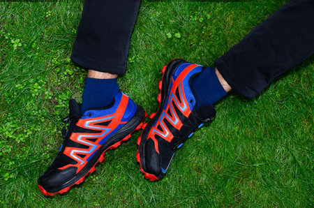 Men's legs shod in a pair of red and blue sneakers are crossed and lie on the green grass.