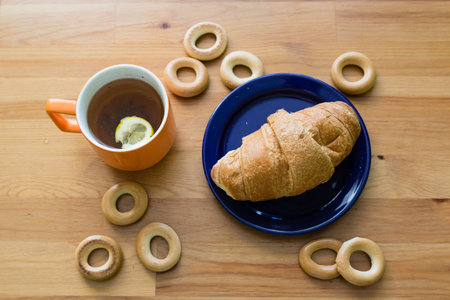 Mug full of black tea with lemon,  croissant on plate and small bagels on wooden tableの写真素材