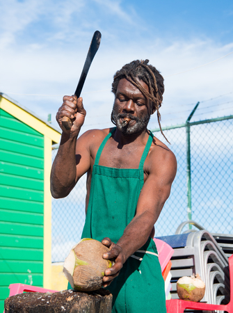 Nassau, Bahamas - January 07, 2016: african american muscular man break big fresh raw coconut with sharp machete smoking cigar outdoor in green apronのeditorial素材