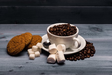 Roasted coffee beans in a white ?up, marshmallows, oatcake, sugar cubes and spoon on vintage wooden background. Side viewの写真素材