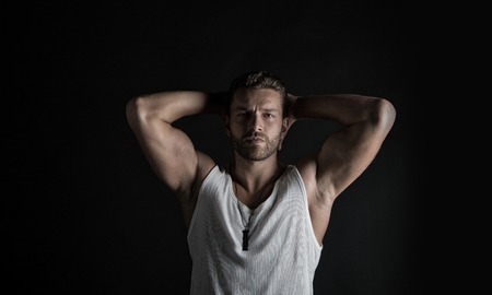 young handsome bearded man macho with muscular hands and beard on serious face in white vest posing in studio on black backgroundの写真素材