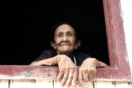 Boca de Valeria, Brazil - December 03, 2015: old mature mulatto woman with kind smiling face holding hands together standing in wooden window brown and white color looking outdoorのeditorial素材