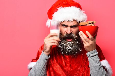 handsome bearded man with stylish mustache and long snowy beard on angry face holding glass of milk with chocolate chip cookies in red santa suit on studio backgroundの写真素材